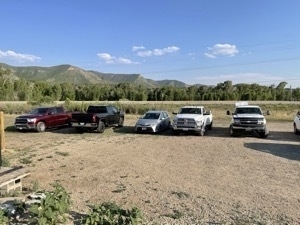 Silver Prius V surrounded by enormous pickup trucks in gravel parking lot.